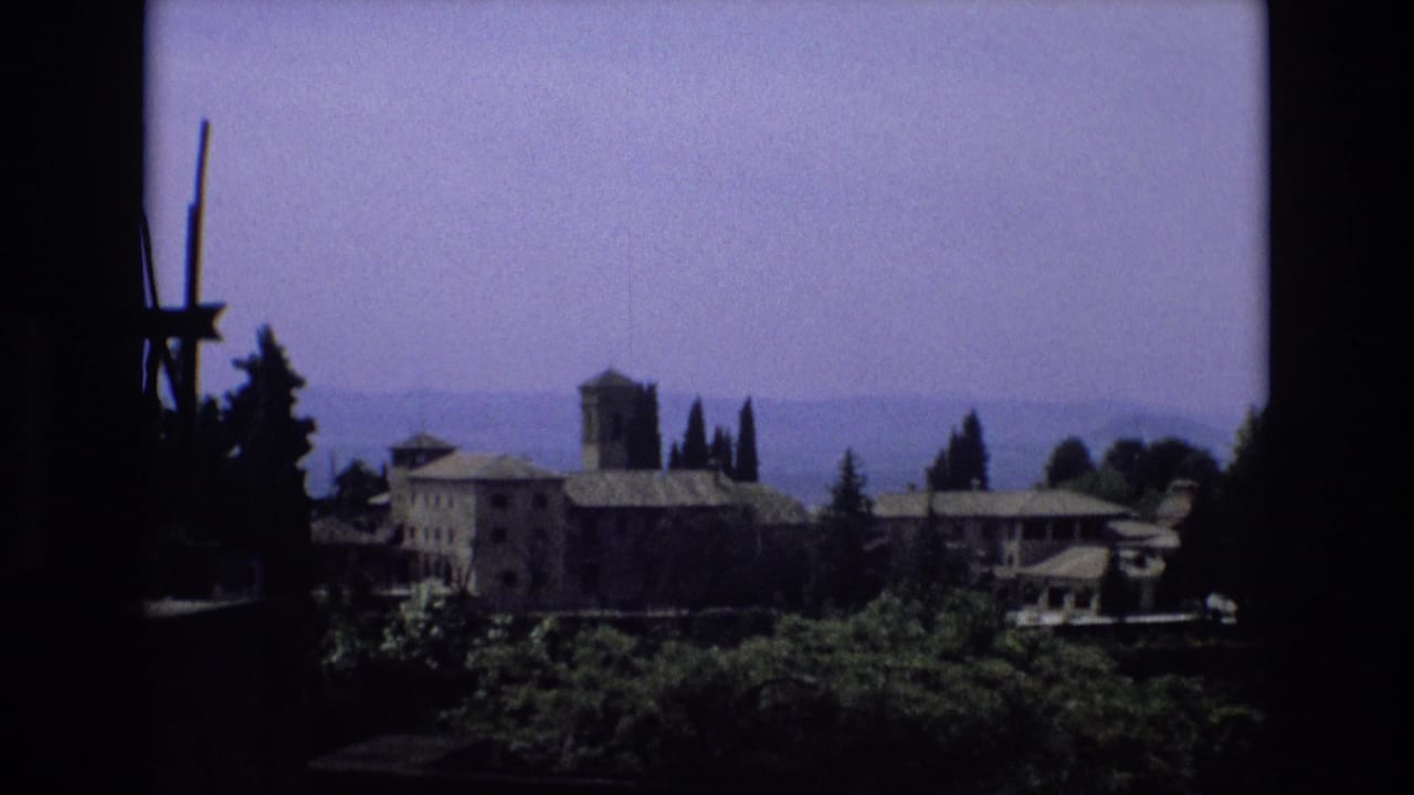 1972 Spain 8mm frame with a towered historic-looking building complex beyond dark foreground framing, cypress-like trees, and hazy hills.