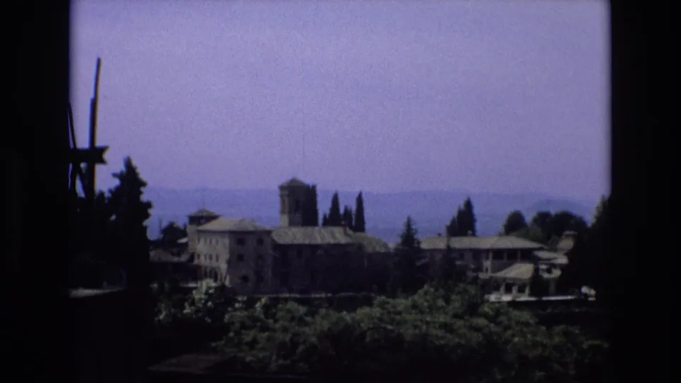 1972 Spain 8mm frame with a towered historic-looking building complex beyond dark foreground framing, cypress-like trees, and hazy hills.
