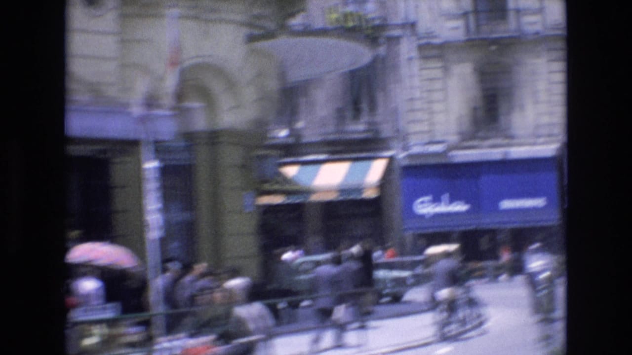 Busy city street with pedestrians, storefront awnings, stone facades, and blurred traffic.