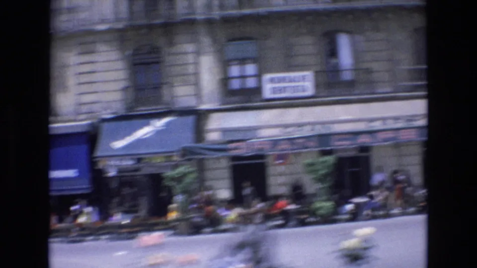A 1972 Spain street frame shows people at or near outdoor cafe seating under awnings, with upper-story windows, signs, and blurred scooters or small motorcycles in the foreground.