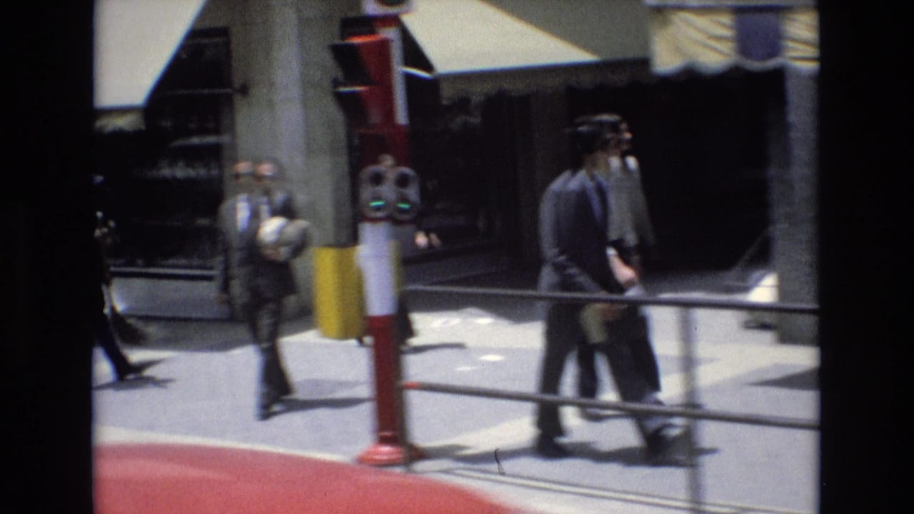 Street-level 8mm frame from Spain in 1972 showing pedestrians near a storefront, a red-and-white street fixture, awnings, and a bright sidewalk.