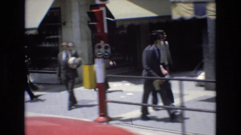 Street-level 8mm frame from Spain in 1972 showing pedestrians near a storefront, a red-and-white street fixture, awnings, and a bright sidewalk.