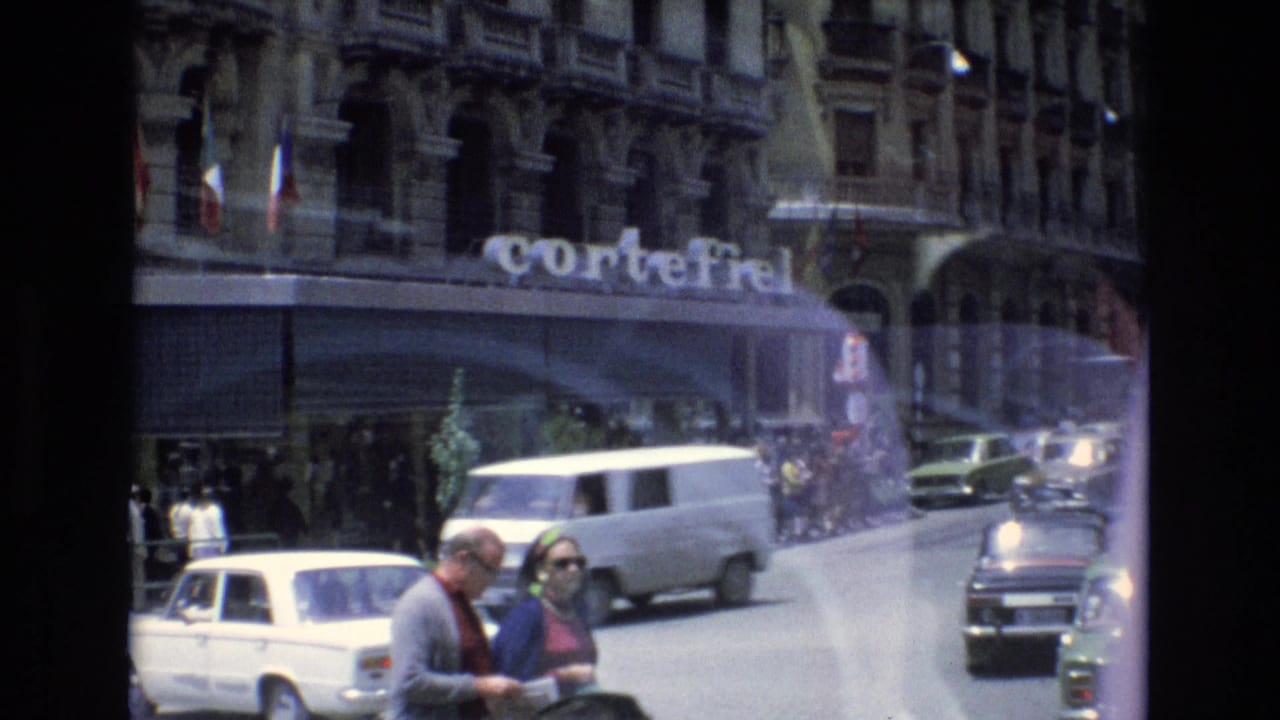 Traffic passes a city building with a CorteFiel sign while pedestrians stand near the foreground.