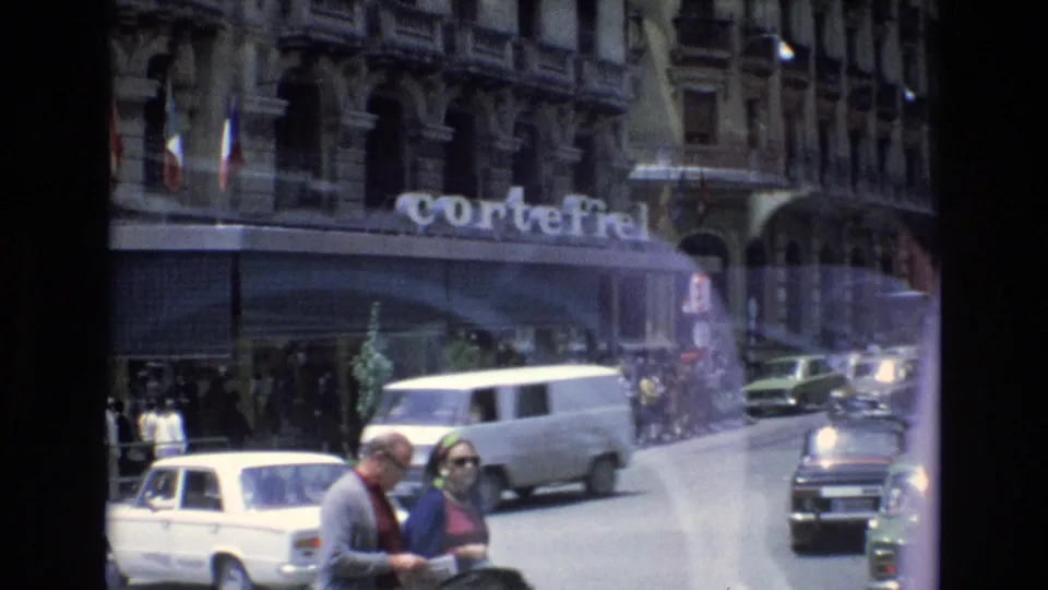 Spain 1972 urban street frame with a visible CorteFiel sign, a white van, compact cars, flags on the building facade, and pedestrians in the foreground.