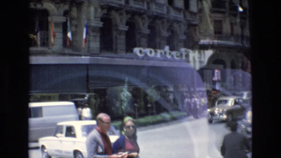 Spain 1972 street frame showing an older man and woman in the foreground, compact cars, a white van, and the CorteFiel sign on a large commercial building.