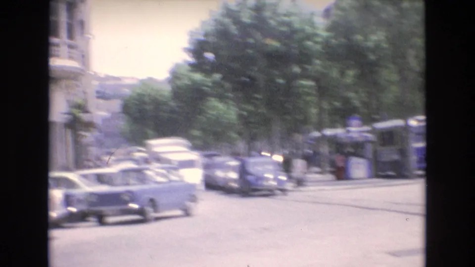 Spain 1972 handheld street view with compact cars, a tree-lined sidewalk, pedestrians, storefront awnings, and soft motion blur from passing traffic.