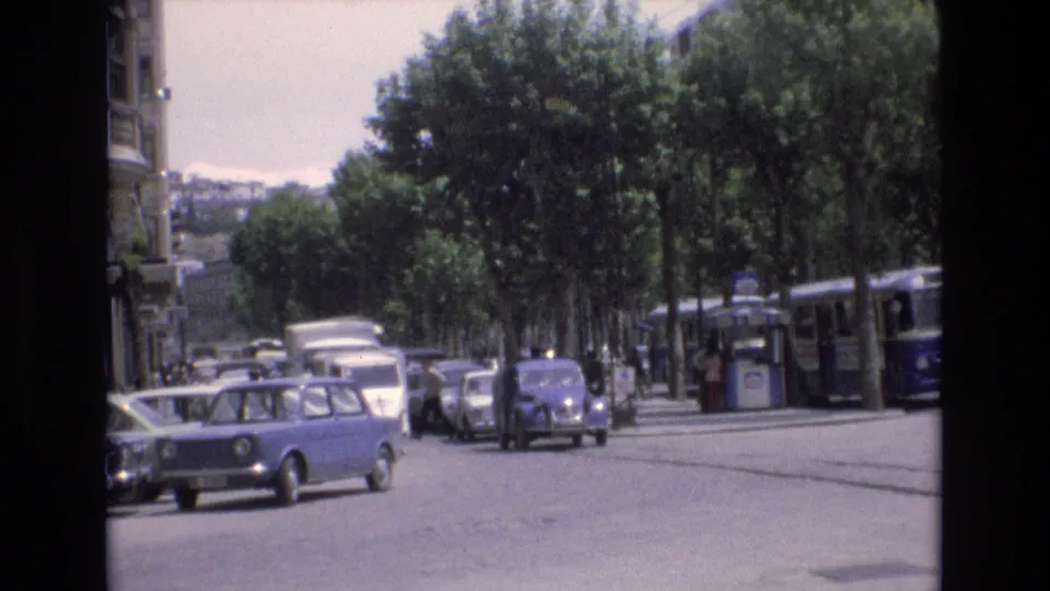 In 1972 Spain, a blue compact car turns across a broad tree-lined city street while other period vehicles and a transit vehicle appear in the background.