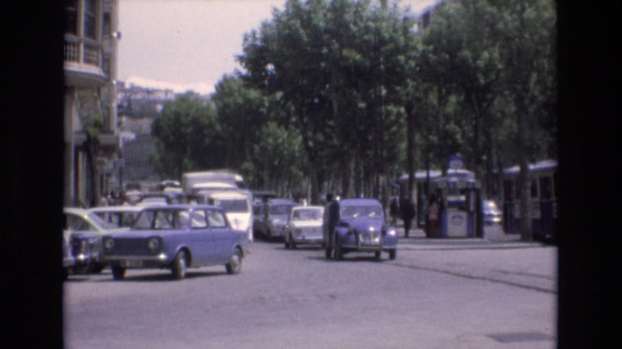 Blue compact car crosses the street as other cars and a transit vehicle line the roadway.