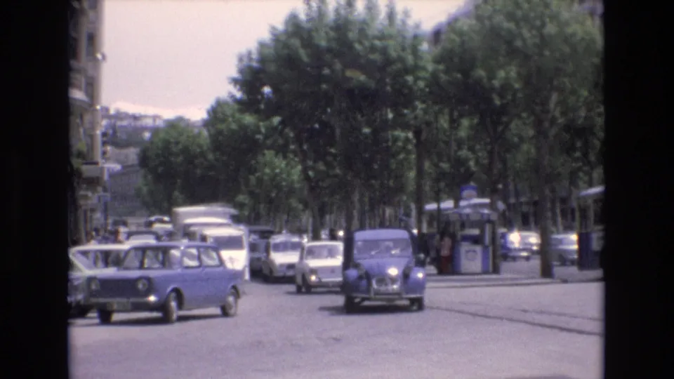 A blue compact car exits the turn while a rounded gray car approaches through traffic on a tree-lined street in Spain, with pedestrians and a kiosk faintly visible near the sidewalk.
