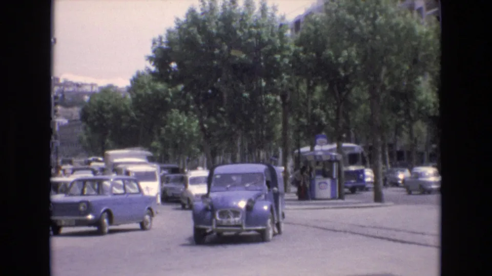 A rounded gray car drives toward the camera on a busy Spanish city street in 1972 as the blue compact car moves away and a blue and white transit vehicle remains near the curb.