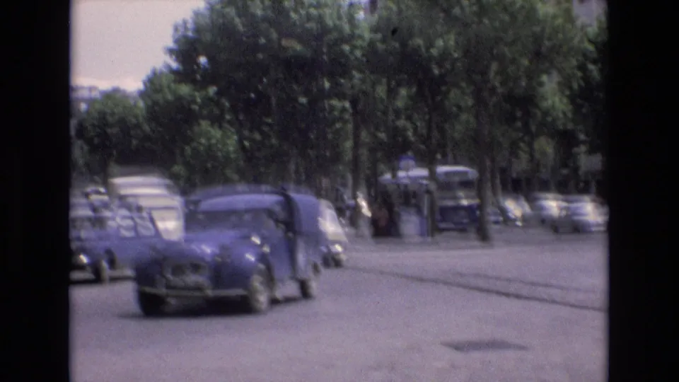 1972 Spain home-movie frame showing a shaded road bordered by trees and parked cars, with a rounded compact car moving through the foreground.