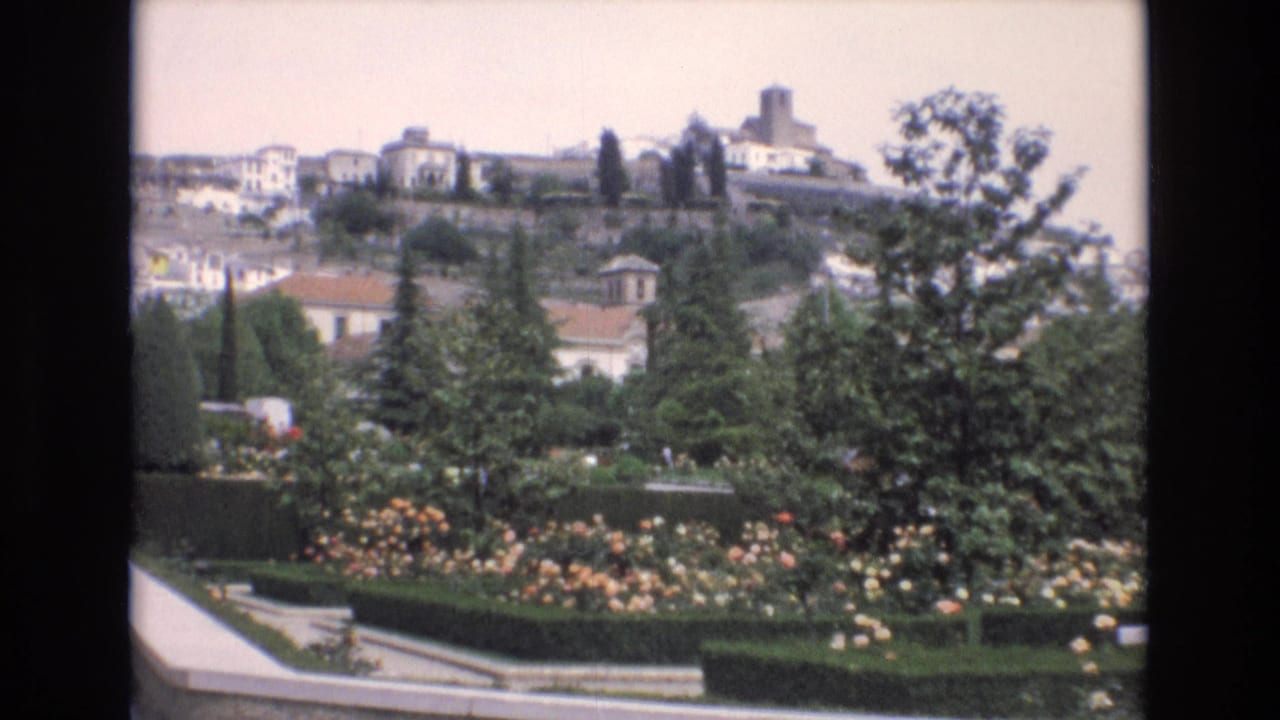 Formal garden with trimmed hedges, rose bushes, paths, trees, and hillside buildings beyond.