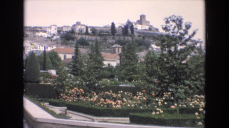 1972 Spain home-movie frame holding on a landscaped garden, with rose beds and clipped greenery in front of hillside buildings and a prominent tower.