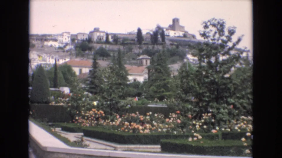 1972 Spain frame showing a formal garden of roses, clipped hedges, paths, trees, and hillside buildings, with a tower visible on the skyline.