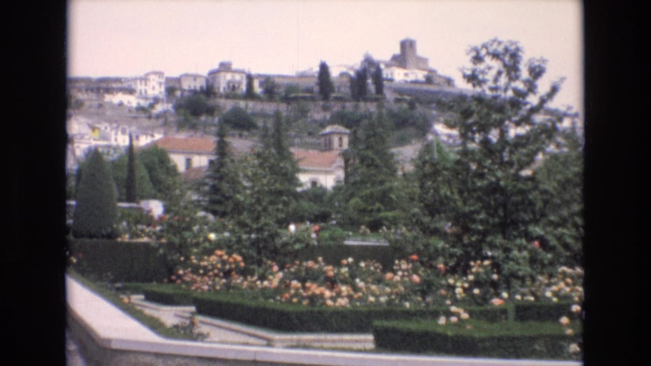 1972 Spain 8mm frame showing a formal public garden with flowering beds, clipped hedges, trees, and white hillside buildings beyond.
