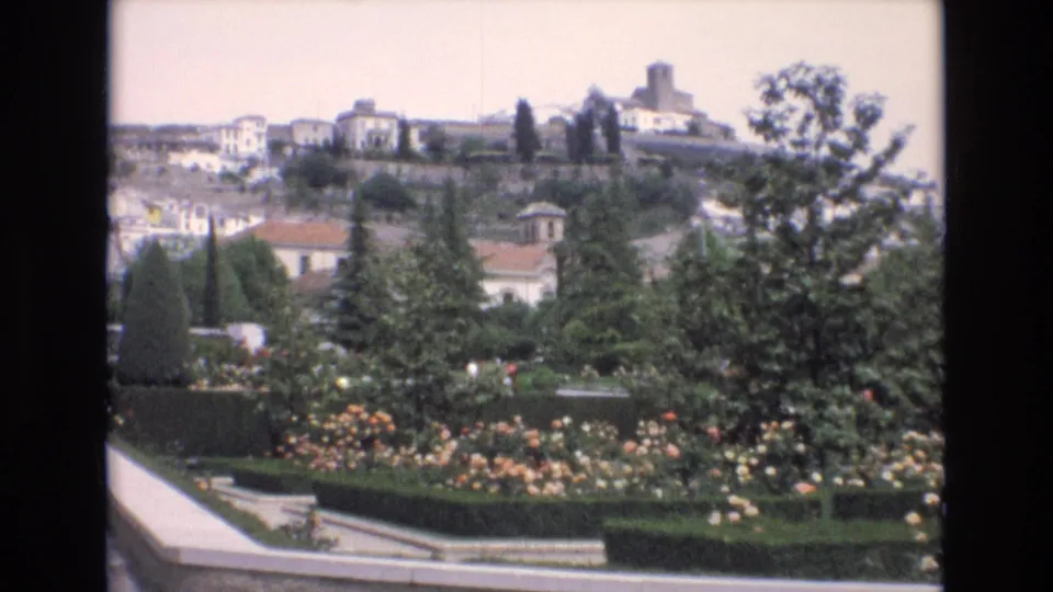 1972 Spain 8mm frame showing a formal public garden with flowering beds, clipped hedges, trees, and white hillside buildings beyond.