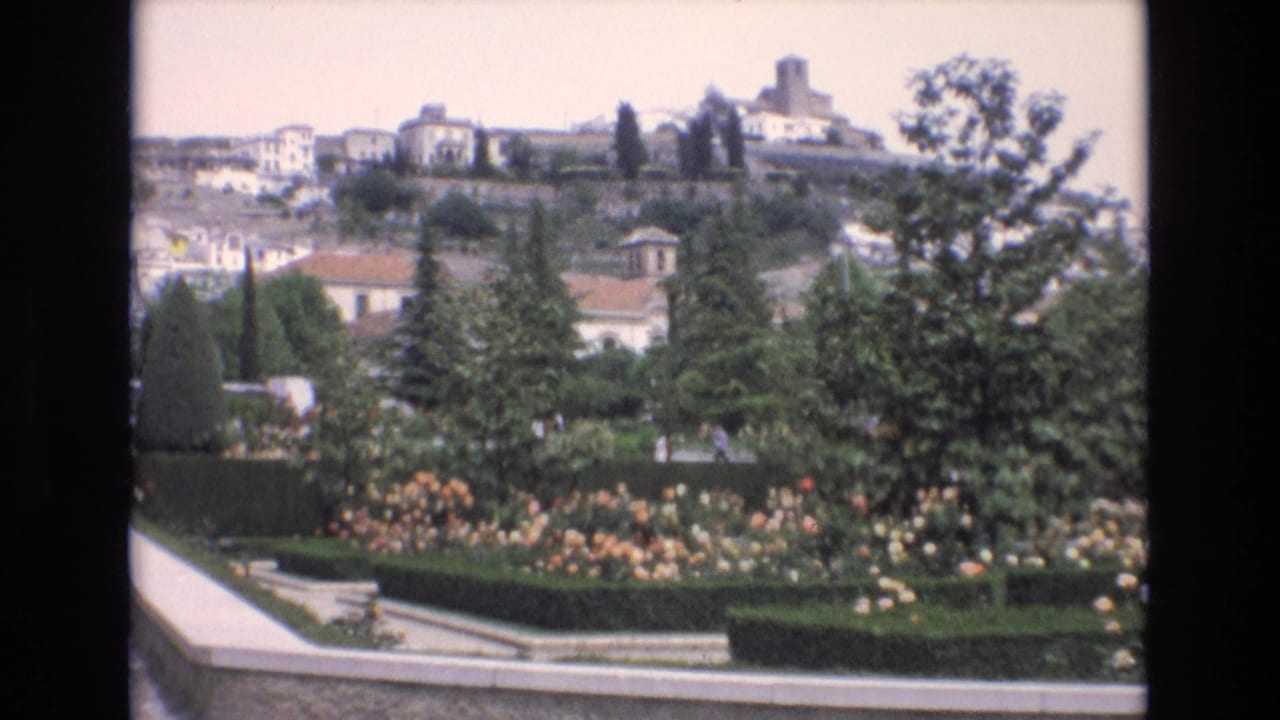 1972 Spain home-movie frame looking across a landscaped garden toward white buildings and a prominent tower on the hillside.
