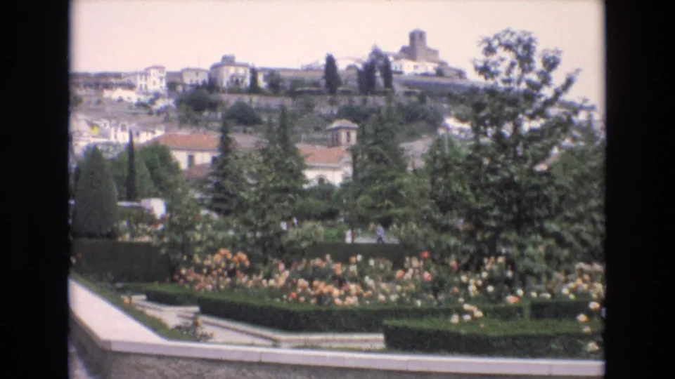 1972 Spain home-movie frame looking across a landscaped garden toward white buildings and a prominent tower on the hillside.