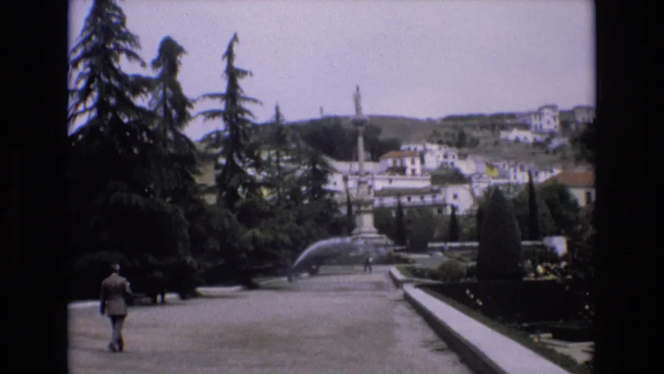 1972 Spain 8mm frame of a wide public-garden walkway with a distant pedestrian, fountain spray, tall trees, and hillside buildings.