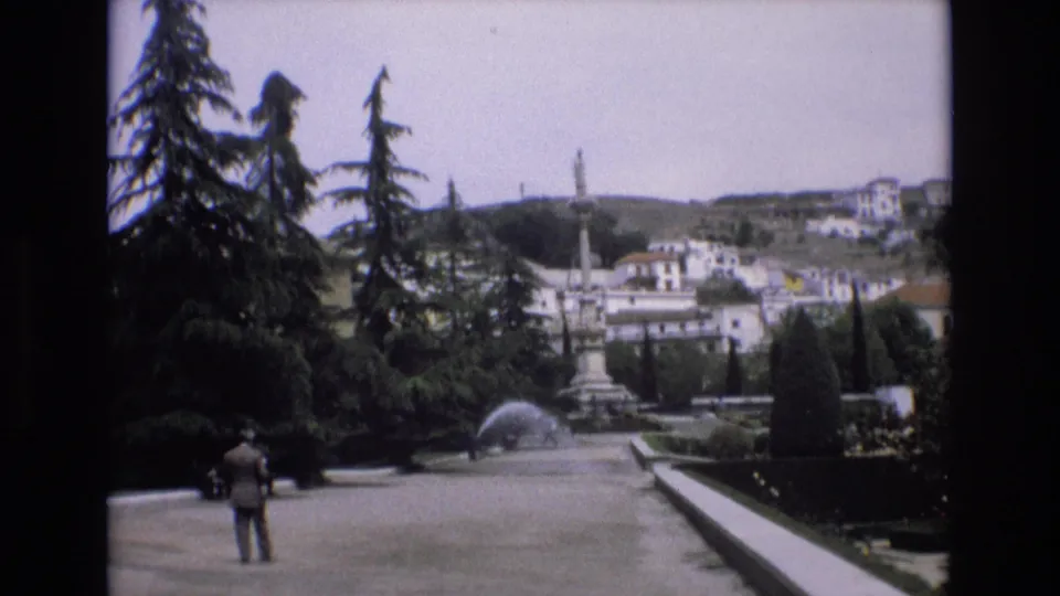 1972 Spain home-movie frame with two distant visitors on a broad garden path, a fountain to the side, tall trees, and town buildings beyond.
