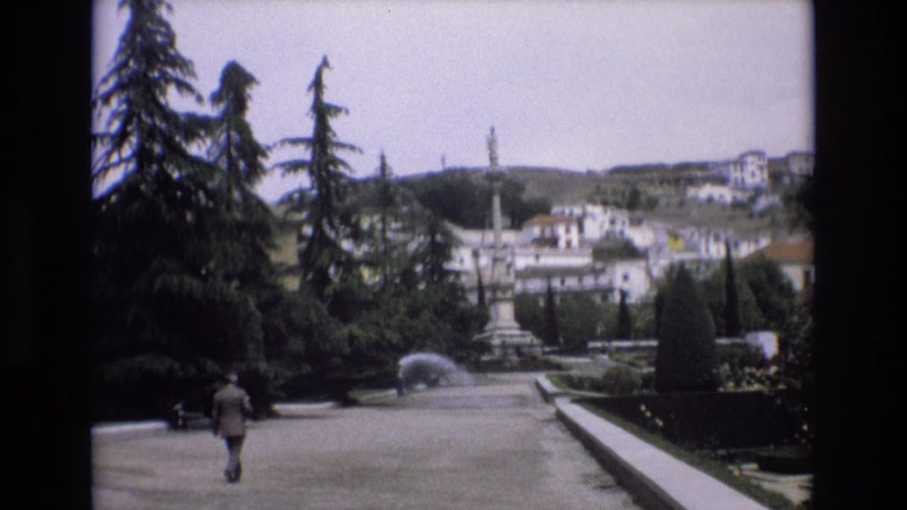 1972 Spain 8mm frame showing a suited man walking away along a broad formal garden path, with evergreen trees, hedges, fountain spray, and hillside buildings beyond.