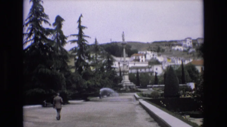 1972 Spain 8mm frame showing a suited man walking away along a broad formal garden path, with evergreen trees, hedges, fountain spray, and hillside buildings beyond.