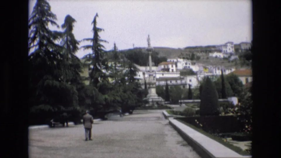 1972 Spain 8mm still showing the suited man farther along the path, with dark evergreen trees, benches, clipped planting beds, fountain spray, and hillside architecture in the background.