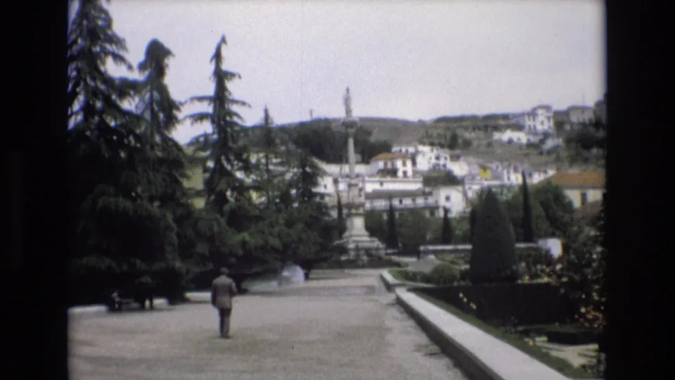1972 Spain frame from 8mm footage showing a solitary suited man walking away through a manicured garden, with a fountain, stone monument, tall evergreens, and white buildings beyond.