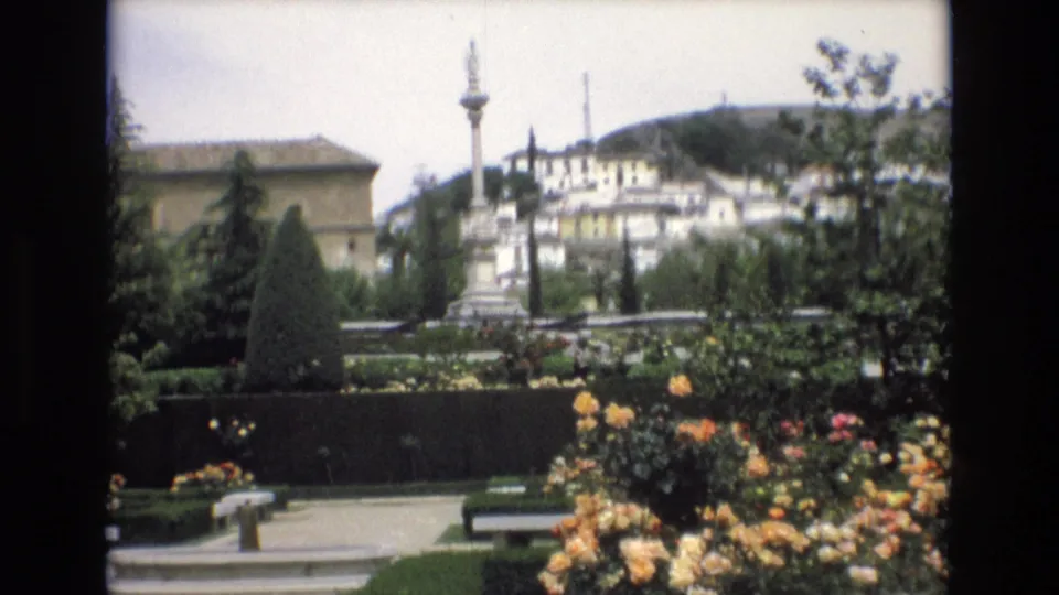 Spain 1972 8mm frame showing a formal garden with blooming roses, clipped hedges, benches, a stone monument, and white hillside buildings in the background.