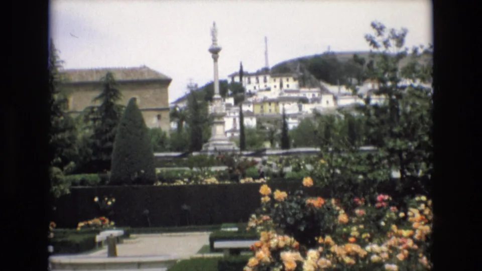 Spain 1972 8mm frame of a landscaped public garden with roses, benches, clipped greenery, a central stone monument, and white buildings on a low hill.