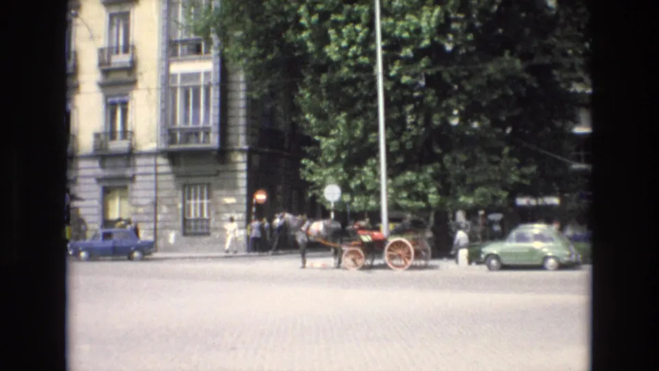 Spain 1972 street view showing pedestrians near a horse-drawn carriage, compact parked cars, leafy trees, signs, and a large stone building facade.