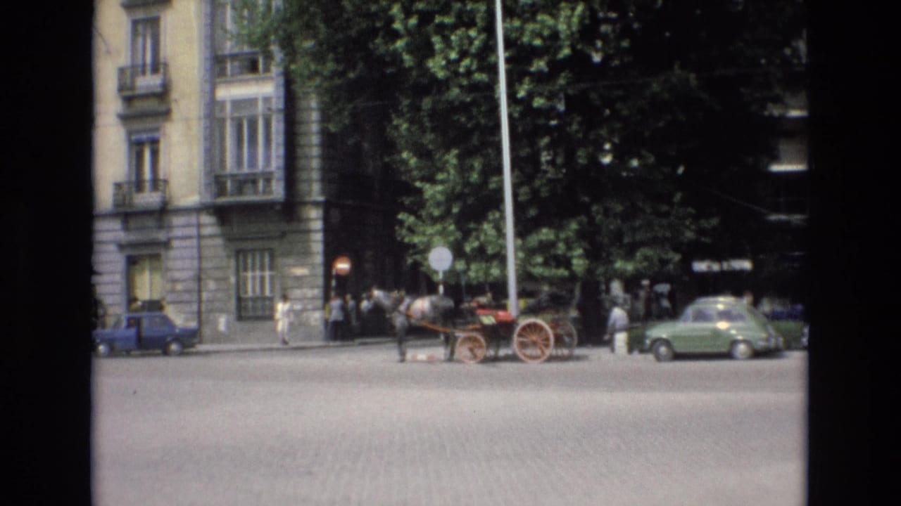 Spain 1972 home-movie frame showing a horse-drawn carriage attraction on a cobblestone street, with pedestrians, trees, stone facades, and parked cars nearby.