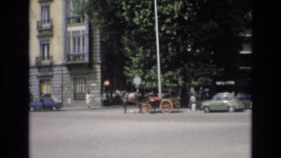 Spain 1972 home-movie frame showing a horse-drawn carriage attraction on a cobblestone street, with pedestrians, trees, stone facades, and parked cars nearby.