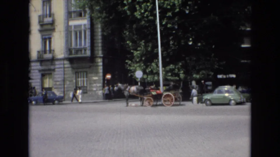 Spain 1972 street view with a horse and red carriage beside parked cars, pedestrians, leafy trees, and multi-story stone architecture.