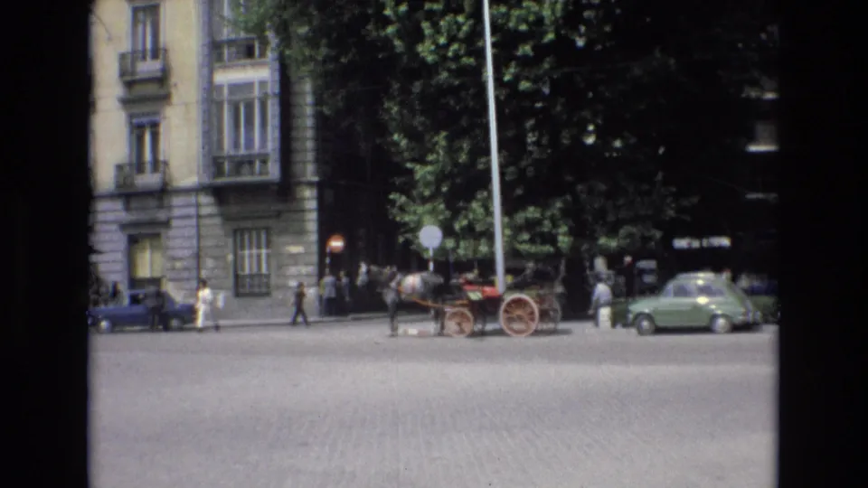 Spain 1972 frame showing pedestrians crossing near a horse-drawn carriage, with parked cars and tall stone-fronted buildings in the background.