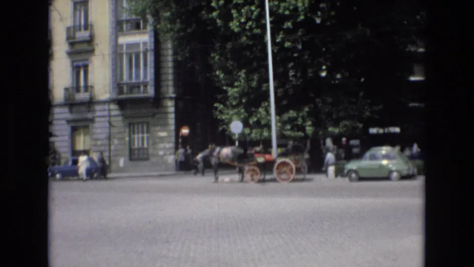 Spain 1972 home-movie still of a stationary horse-drawn carriage on a cobblestone street, with pedestrians, parked cars, trees, and stone urban facades visible.