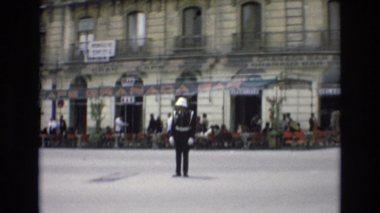 In Spain in 1972, a white-helmeted traffic guard stands in a city street in front of the Granada Bar, with cafe chairs, pedestrians, balconies, and storefront signs behind him.