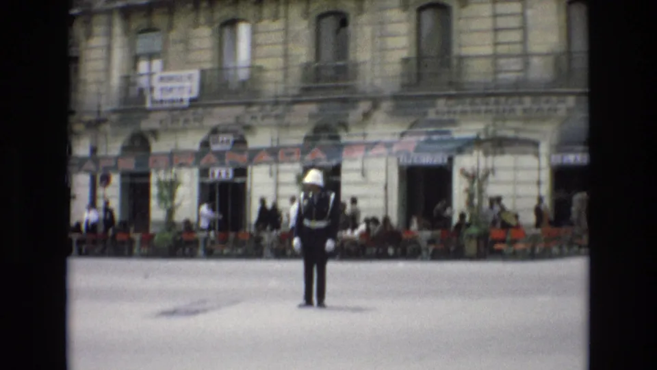 In Spain in 1972, a white-helmeted traffic guard stands in a city street in front of the Granada Bar, with cafe chairs, pedestrians, balconies, and storefront signs behind him.