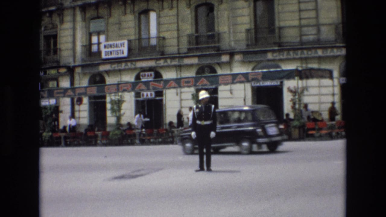 A uniformed traffic guard stands near the center of a Spanish city street as a dark period car passes, with the Granada Bar, cafe tables, pedestrians, and upper-floor balconies visible behind.