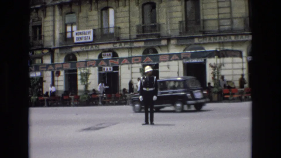 A uniformed traffic guard stands near the center of a Spanish city street as a dark period car passes, with the Granada Bar, cafe tables, pedestrians, and upper-floor balconies visible behind.