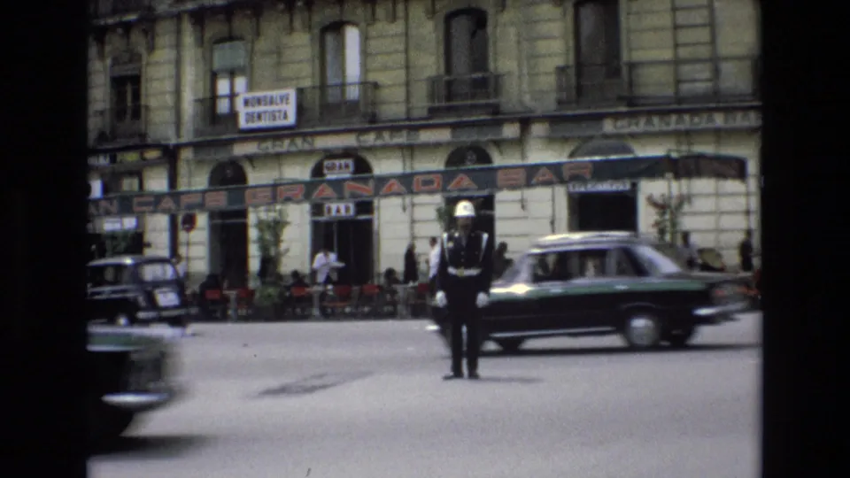 The 1972 Spain street scene shows the traffic guard standing in place while a green sedan and a darker vehicle move through the frame in front of cafe seating and commercial signs.