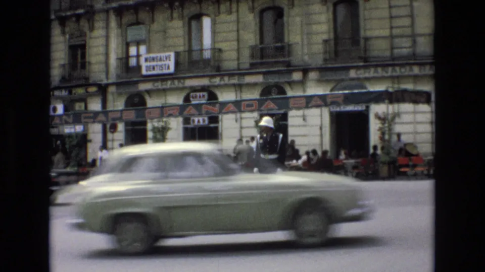 A green period car crosses the foreground in motion blur while the white-helmeted traffic guard remains visible near the Granada Bar and cafe seating in the background.