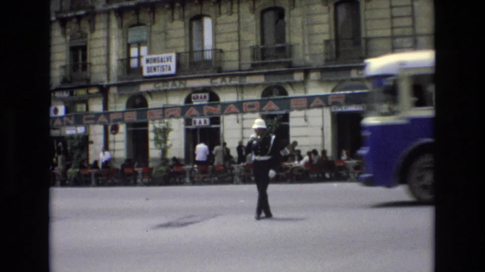 A uniformed traffic officer directs street movement while a blue-and-white public bus passes in front of Granada Bar and nearby shop signs in Spain, 1972.