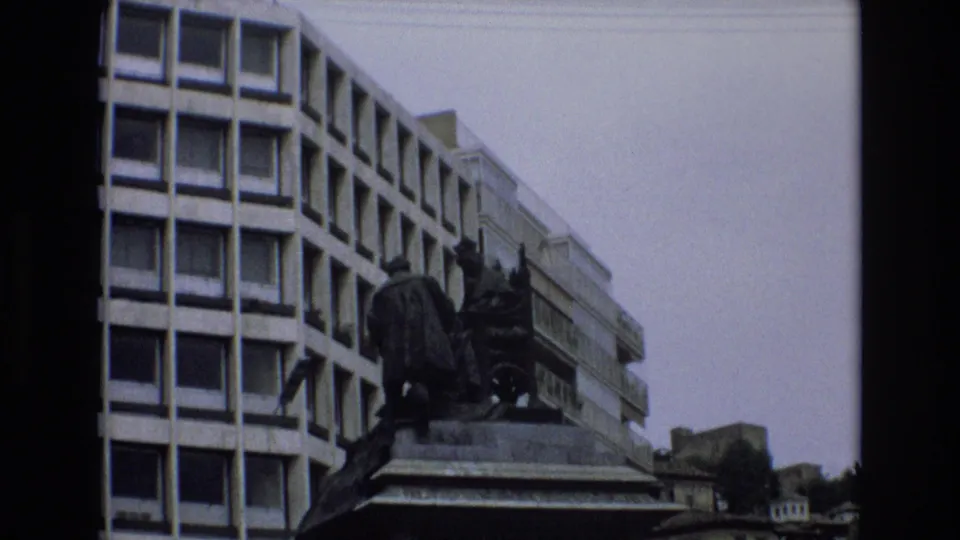 Handheld upward view in Spain, 1972, showing a multi-story modern building beside a dark monument or statue under a pale sky.