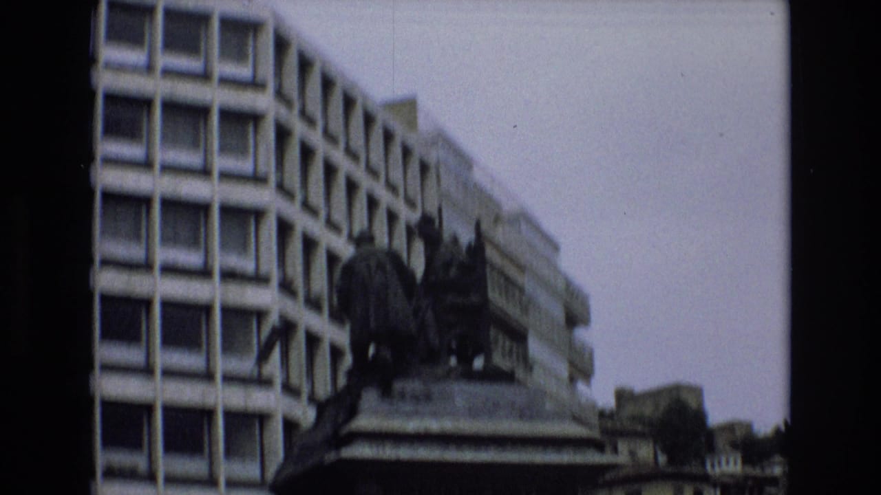 1972 Spain 8mm frame showing a dark bronze monument on a stone base, partly silhouetted against a modern building with repeated square windows.