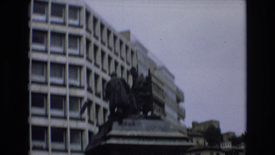 1972 Spain 8mm frame showing a dark bronze monument on a stone base, partly silhouetted against a modern building with repeated square windows.