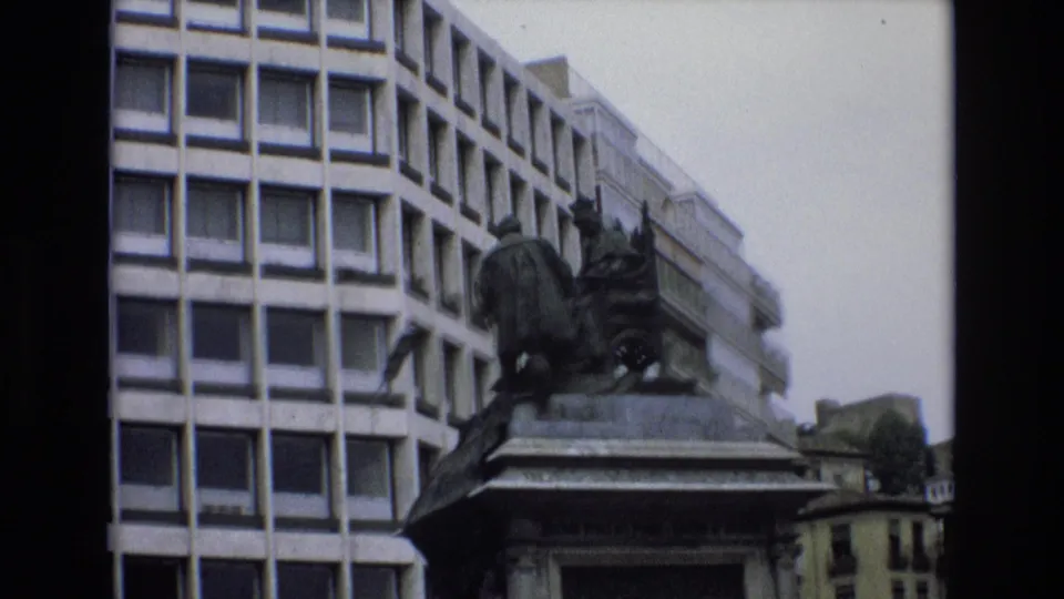 1972 Spain 8mm frame with the monument and stone pedestal more centered, backed by a tall facade of square windows and older buildings at the side.