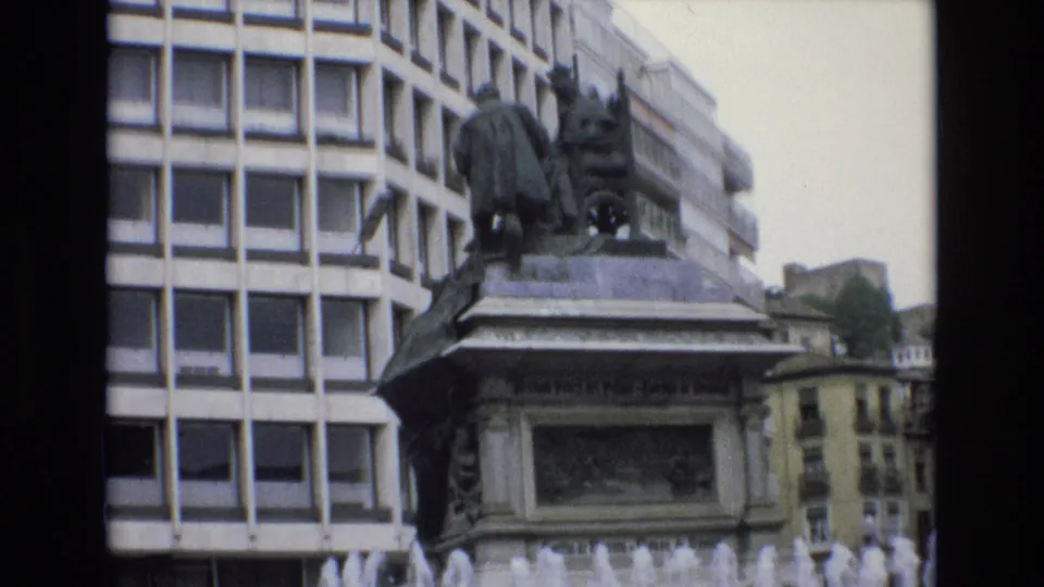 1972 Spain 8mm frame showing the stone pedestal and bronze monument in front of a modern building, with fountain jets rising at the bottom of the view.