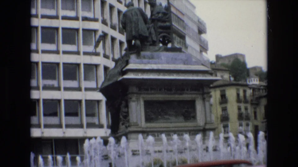 1972 Spain 8mm frame with multiple fountain jets in the foreground, a stone monument pedestal behind them, and a grid-window building filling the background.
