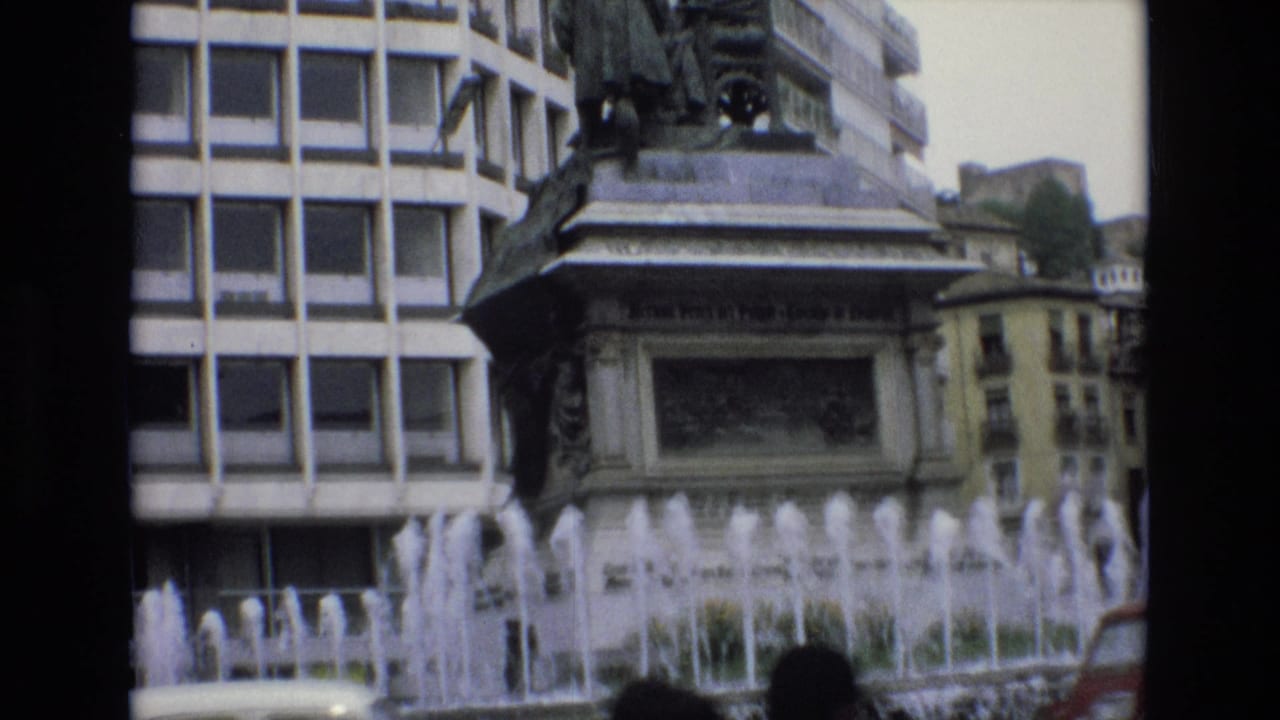 1972 Spain 8mm frame showing a city fountain before an ornate monument base, with a grid-window building and passing street traffic in the foreground.